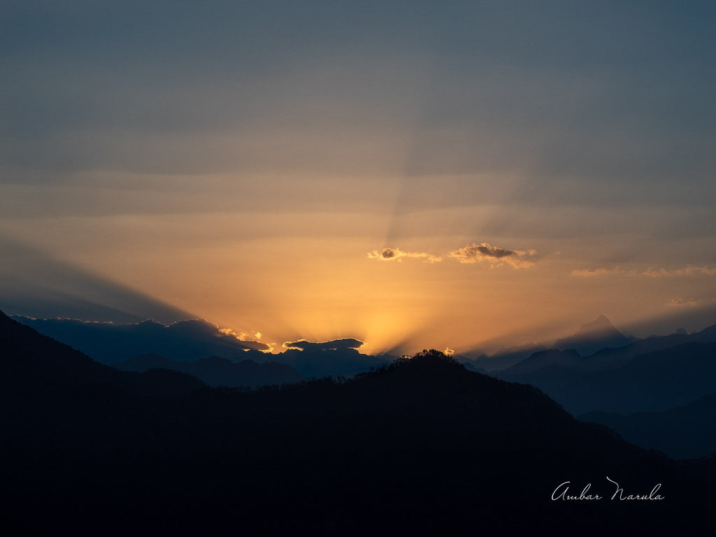 A Landscape shot of the sun rising behind a mountain. Clouds cover the sun, only allowing the rays to escape. The sun is completely hidden from view.